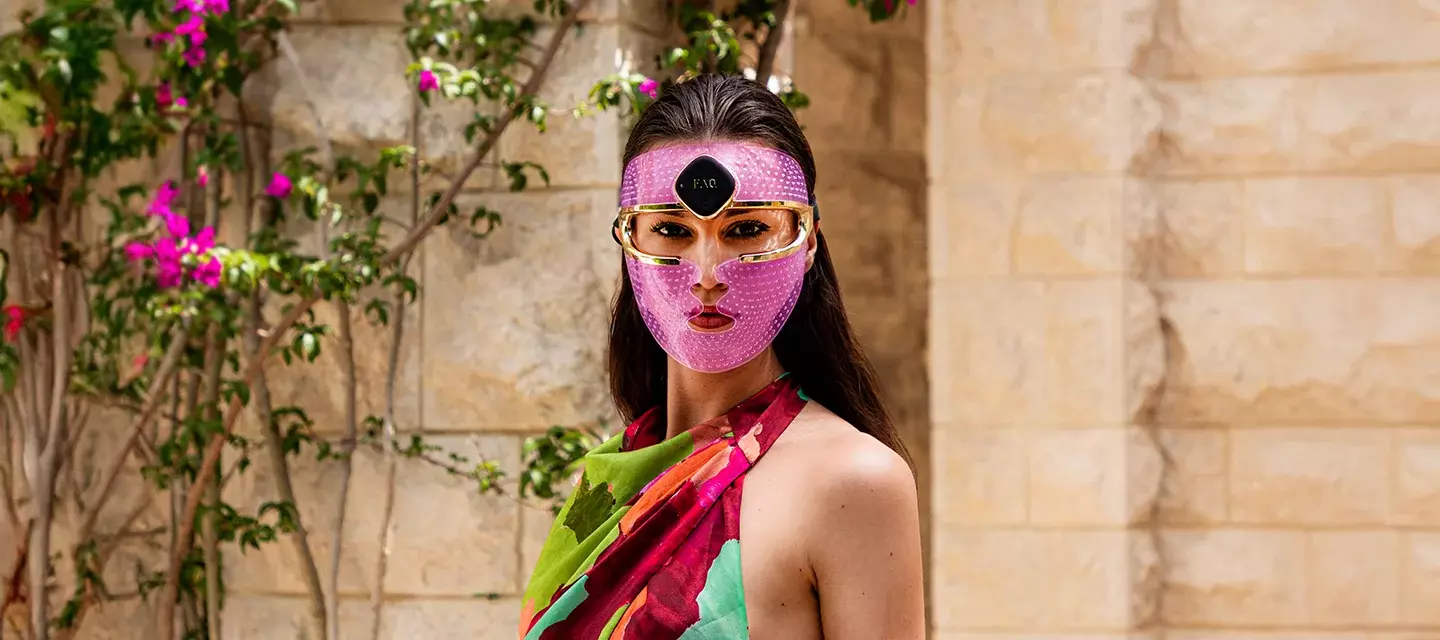 A gorgeous woman with dark hair and dark eyes, in a summer dress, in front of purple flowers, wearing a led face mask set on a purple light 